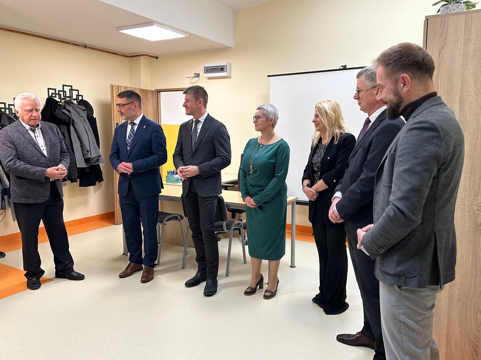 Zdjęcie przedstawia uroczystość otwarcia Klubu Rozwoju Cyfrowego i nowego mieszkania treningowego przy ulicy Wrzesińskiej w Swarzędzu. Na zdjęciu widać zaproszonych gospodarzy i gości stojących we wnętrzu głównej sali w Klubie Rozwoju Cyfrowego. Od lewej stoją: przemawiający Ryszard Dyzma - Przewodniczący Komisji Spraw Społecznych Rady Miejskiej w Swarzędzu, Tomasz Zwoliński - drugi zastępca burmistrza miasta i gminy Swarzędz, Grzegorz Taterka - pierwszy zastępca burmistrza, Agnieszka Maciejowicz - dyrektor Centrum Usług Społecznych w Swarzędzu, Katarzyna Kretkowska - członkini zarządu województwa wielkopolskiego, Marian Szkudlarek - burmistrz miasta i gminy Swarzędz oraz Grzegorz Grygiel - dyrektor Regionalnego Ośrodka Polityki Społecznej w Poznaniu.