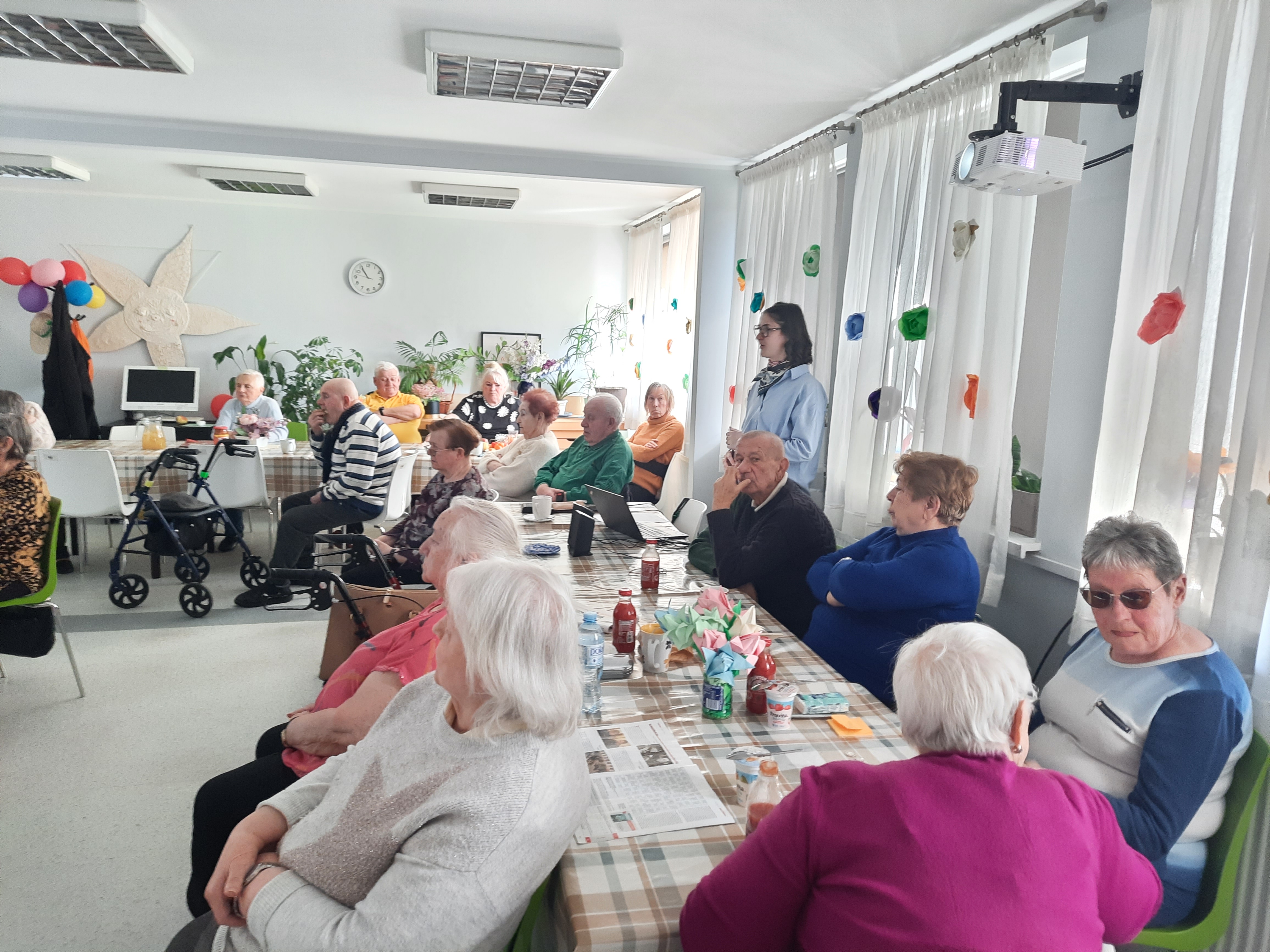 Zdjęcie przedstawia widok głównej sali w Dziennym Domu Senior Wigor oświetlonej słońcem. Seniorzy siedzą przy dwóch długich stołach ustawionych do siebie prostopadle. Pomiędzy siedzącymi i oglądającymi seniorami widać stojącą i opowiadającą Żuliet.