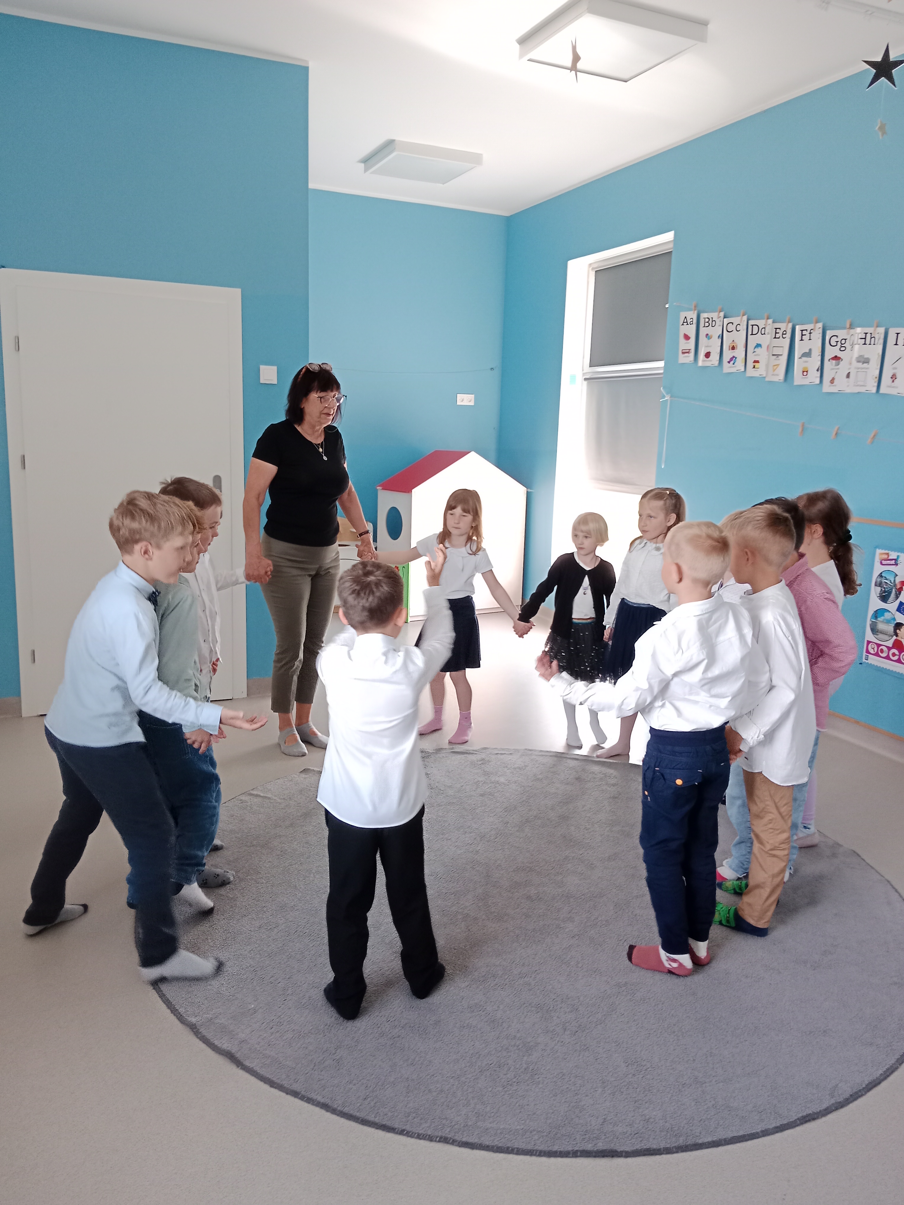 Zdjęcie przedstawia seniorkę oraz grupę dzieci trzymających się za ręce i stojących razem na dywanie w kółeczku.
