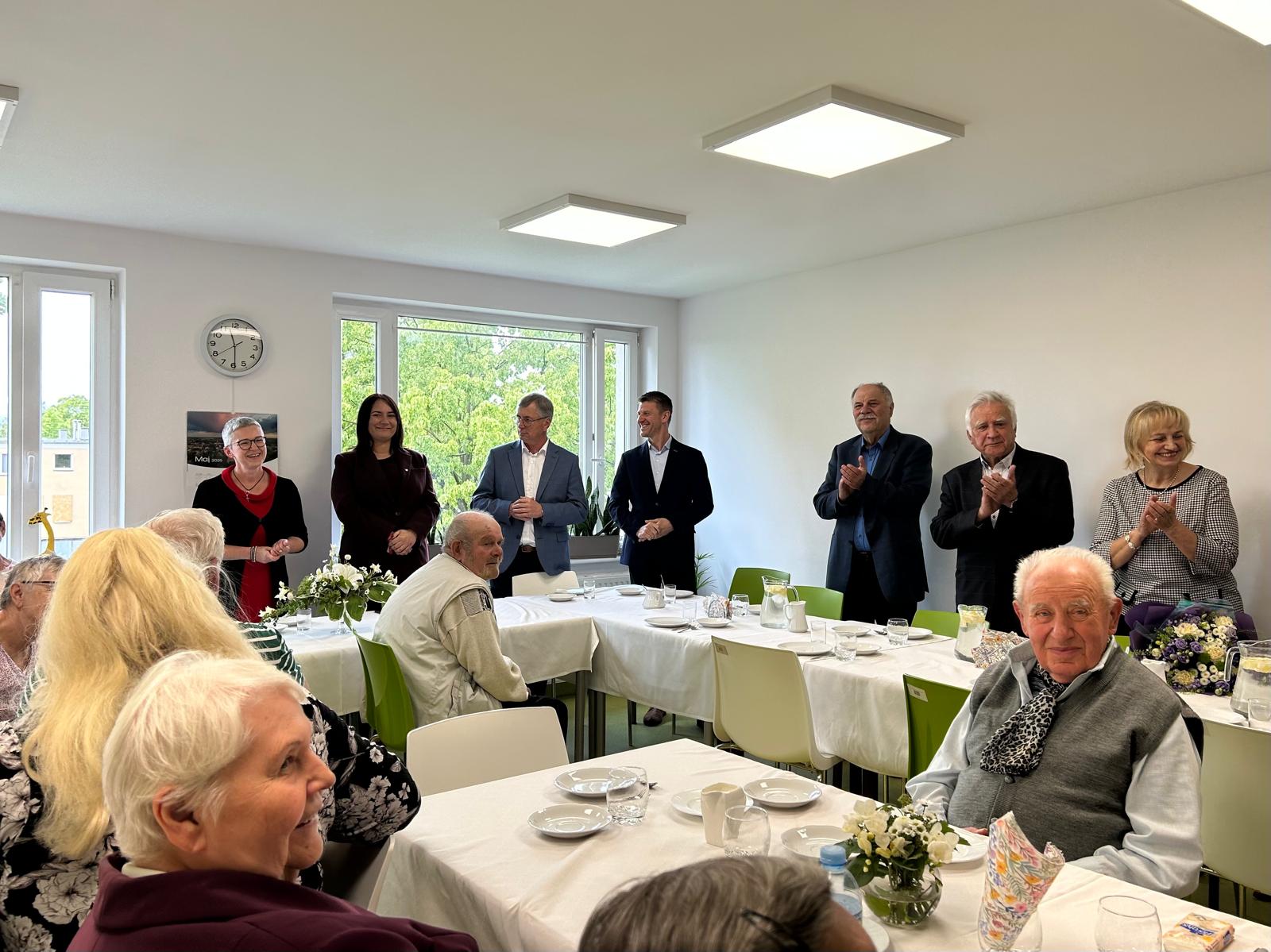 Za uroczystymi stołami siedzą uśmiechnięci seniorzy i seniorki. W tle przy dwóch długich stołach stoją: dyrektor Centrum Usług Społecznych, pierwsza wicewojewoda, burmistrz i jego pierwszy zastępca, przewodnicząca rady miejskiej w Swarzędzu i radni.