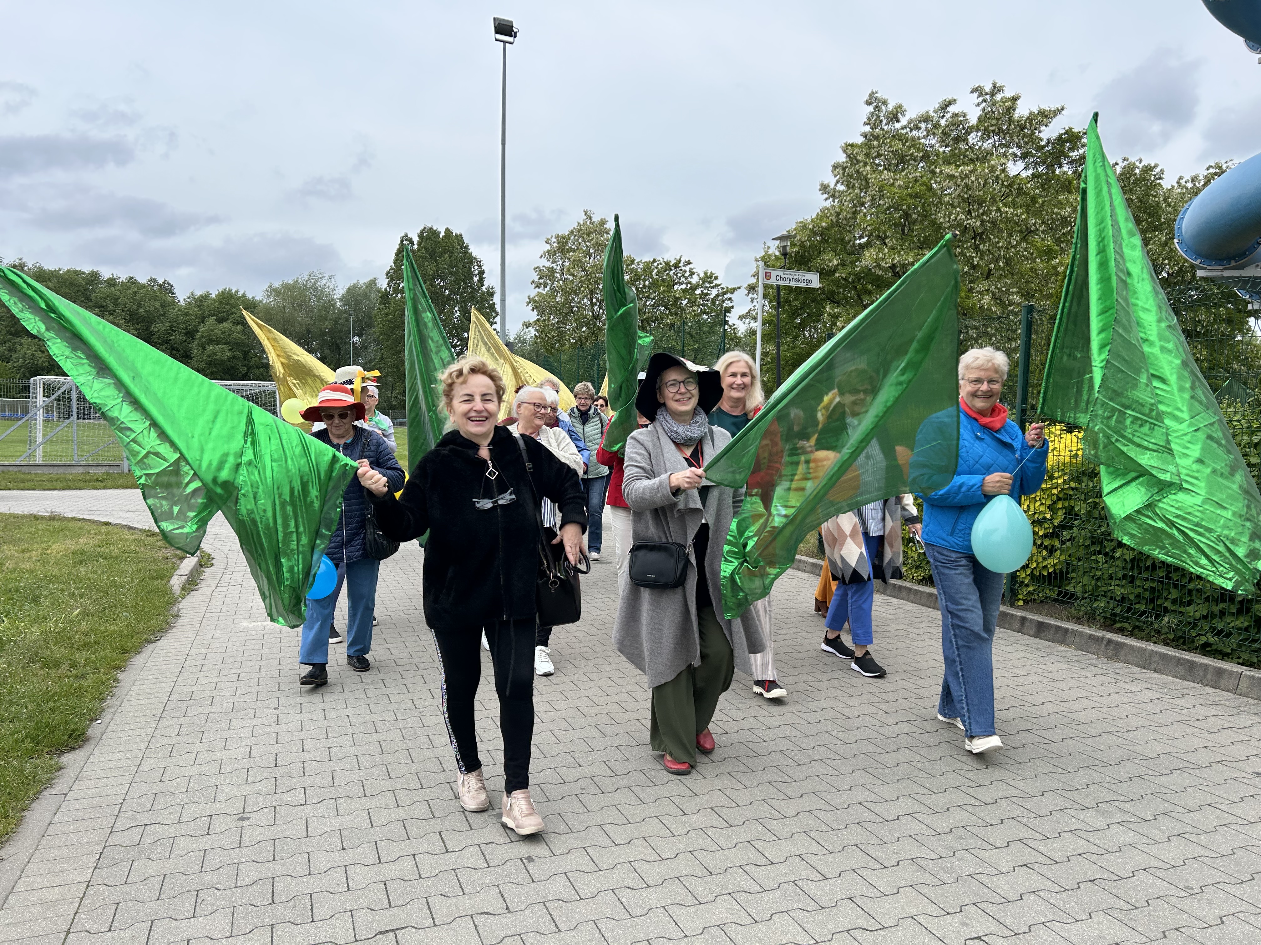 Zdjęcie przedstawia uśmiechniętych uczestników barwnego korowodu rozpoczynającego Senioriadę. Idące z radością osoby trzymają w rękach drzewca, na których powiewają duże flagi z zielonego połyskującego materiału.