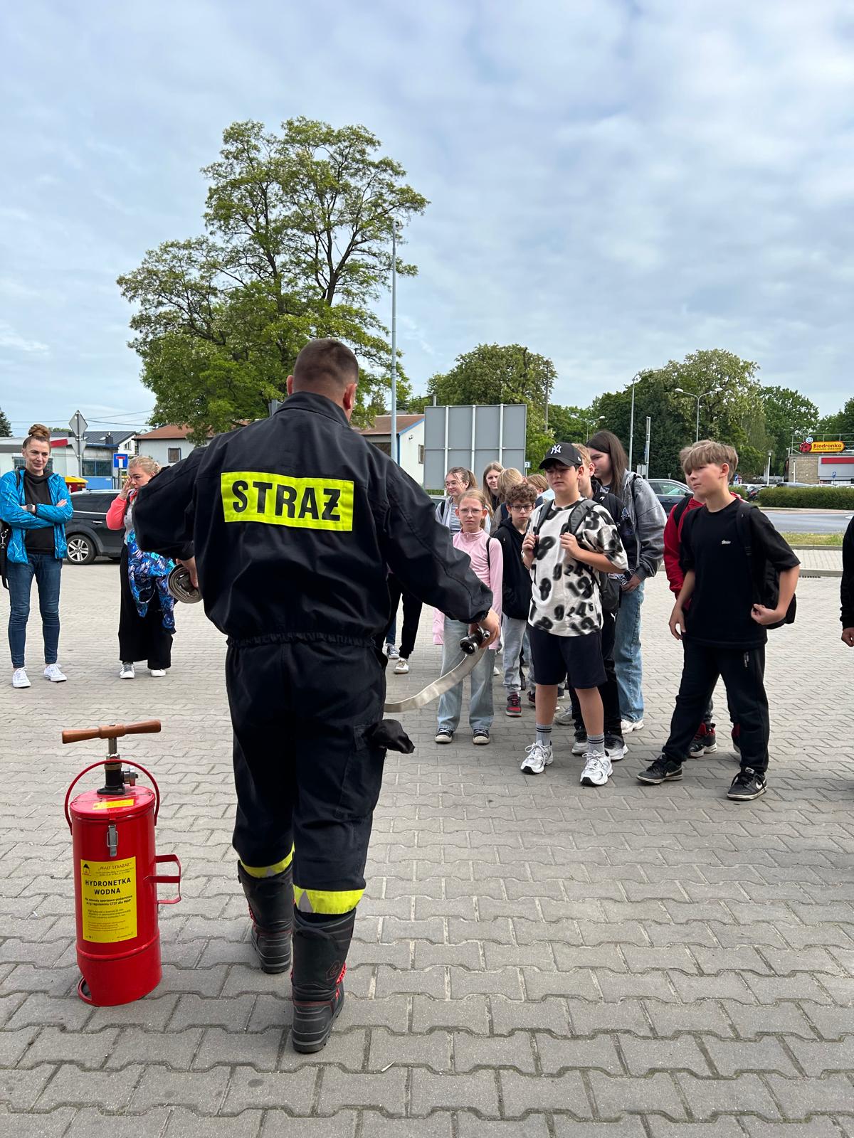 Zdjęcie przedstawia punkt startowy gry miejskiej pod tytułem Ratujemy bo umiemy. Na pierwszym planie widać strażaka tłumaczącego grupie uczniów konkurencję, do której za chwilę przystąpią. Obok strażaka stoi czerwona hydronetka wodna z żółtą etykietą.
