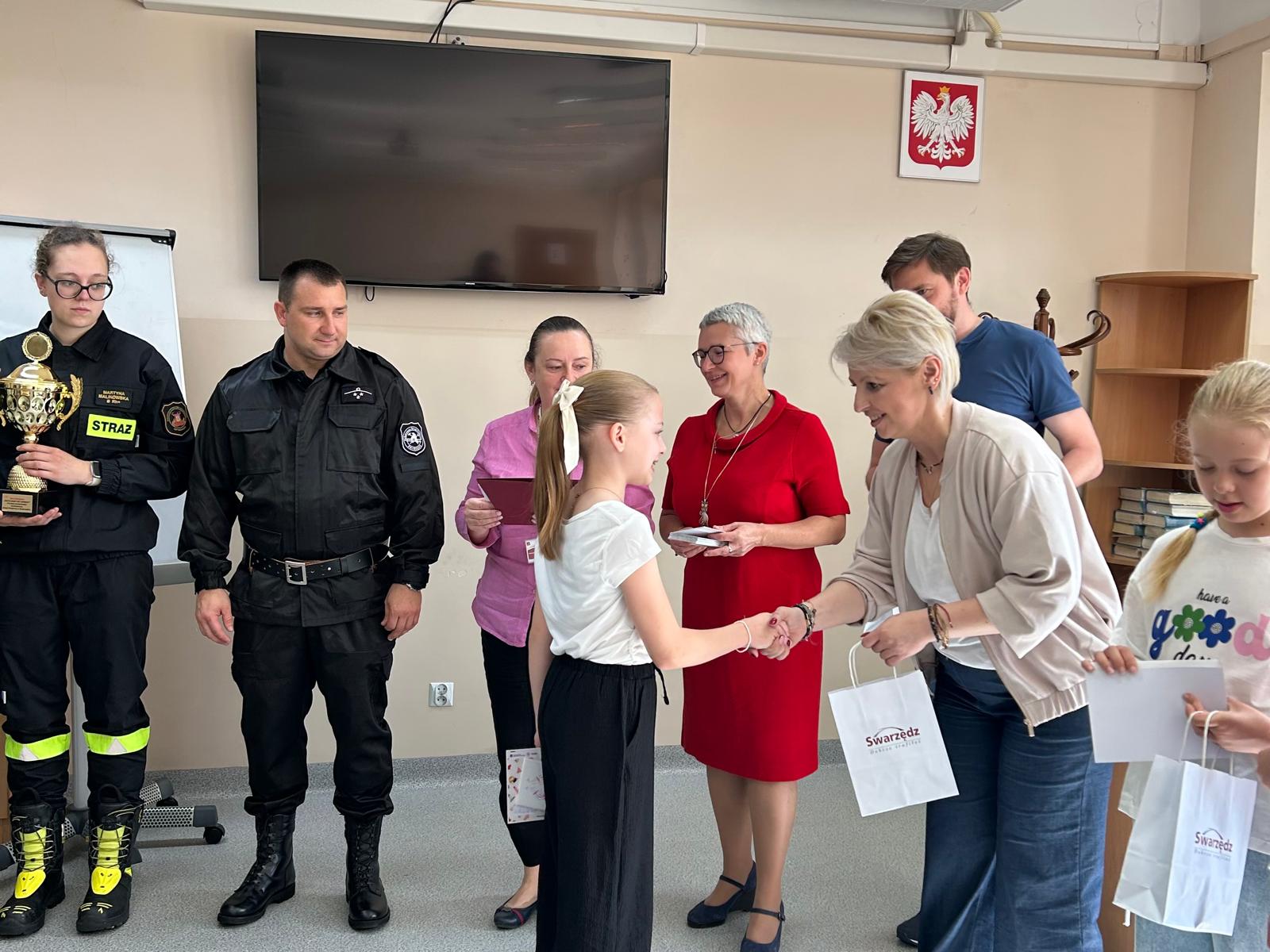 Zdjęcie ukazuje moment wręczenia nagrody i dyplomu jednej z zawodniczek przez dyrektor szkoły podstawowej numer dwa w Swarzędzu. Po prawej stronie dyrektor stoi troje przedstawicieli Centrum Usług Społecznych oraz dwoje strażaków.