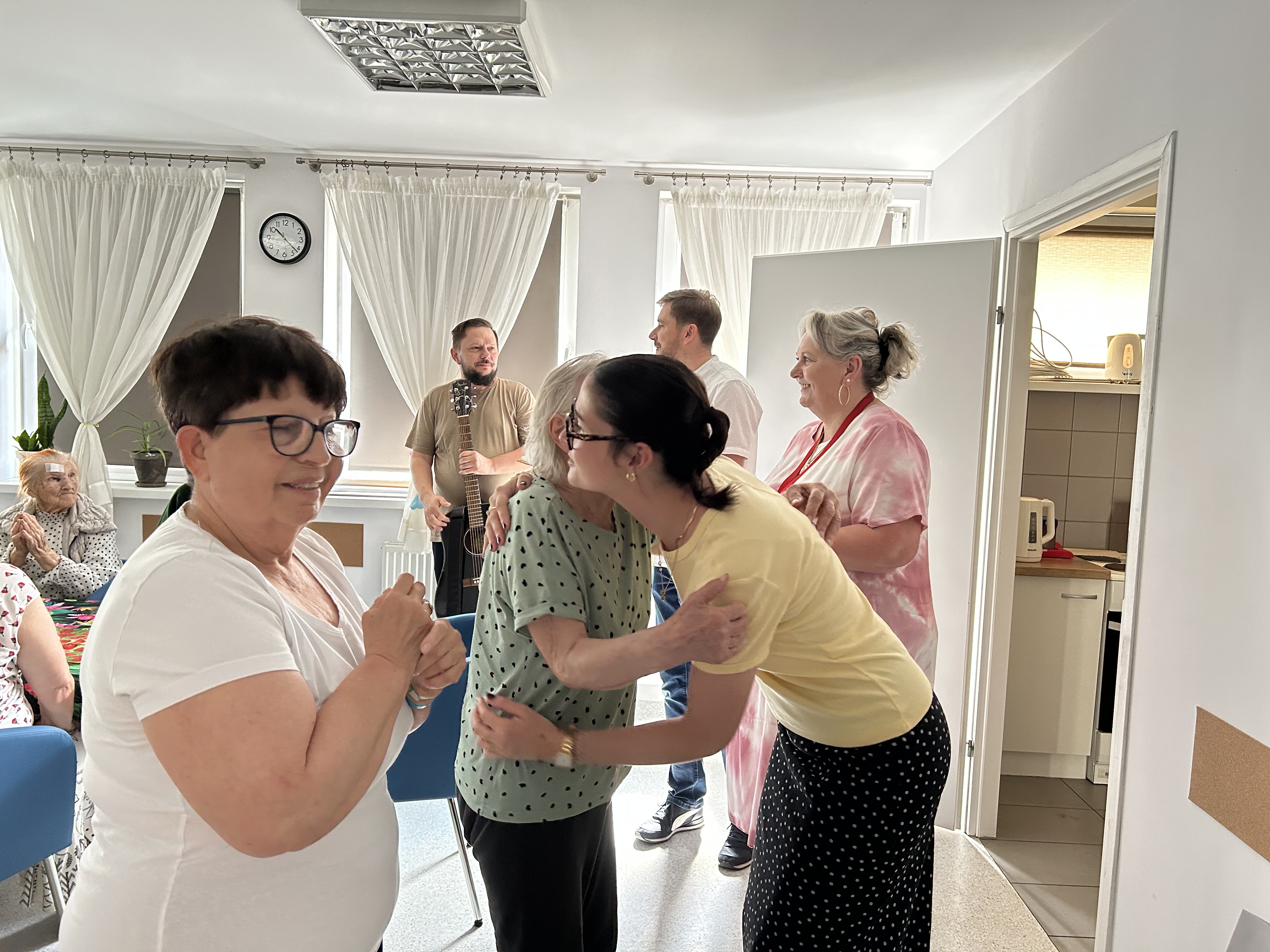Wnętrze Dziennego Domu Pomocy. Pożegnanie Żuljet Żama wolontariuszki Europejskiego Korpusu Solidarności. Wolontariuszka przytula się do seniorki składającej jej życzenia. Druga seniorka czeka w kolejce do Żuljet. W tle widać troje pracowników.