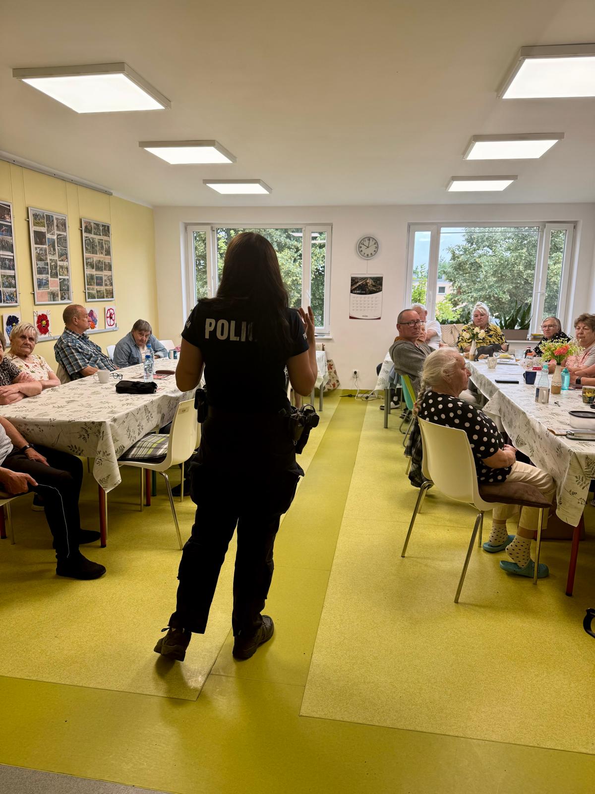 Zdjęcie przedstawia grupę seniorów siedzących przy stolikach zwróconych do policjantki, która stoi na środku pomieszczenia.