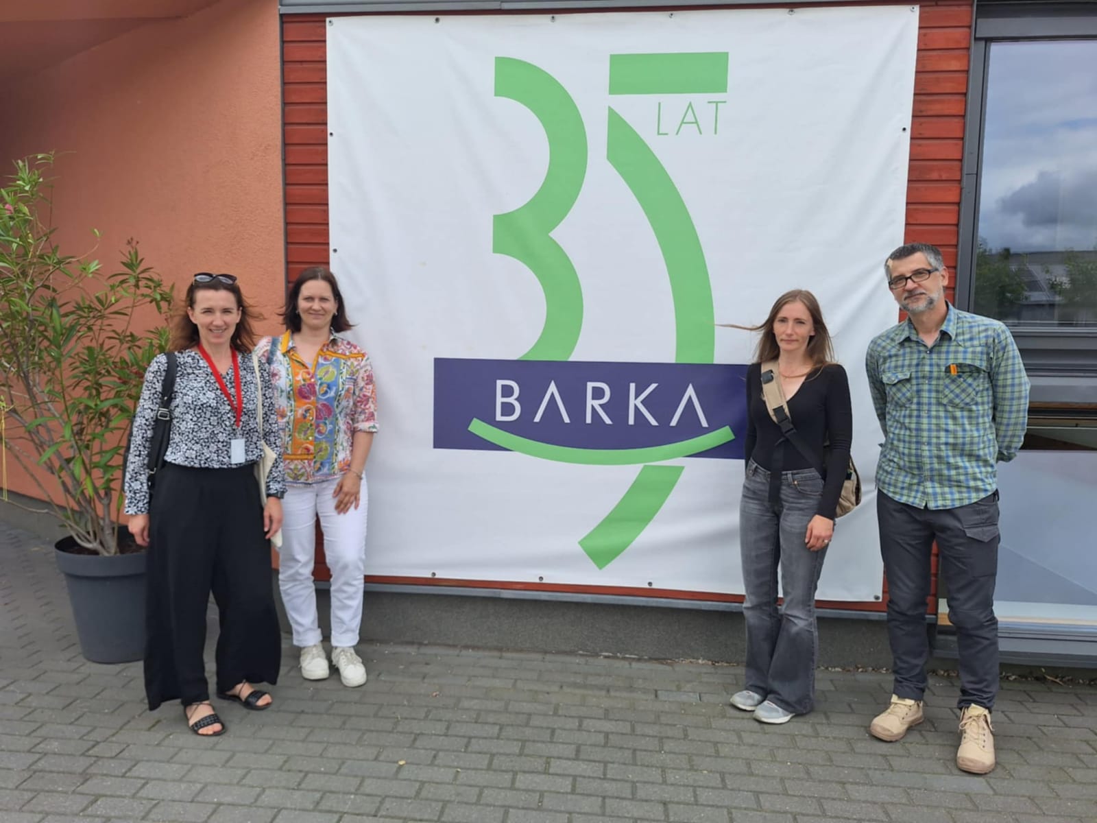 Czworo uczestników spotkania stoi na tle banera z napisem Barka 35 lat. Baner wisi na ścianie budynku, który jest siedzibą Fundacji Barka w Poznaniu.