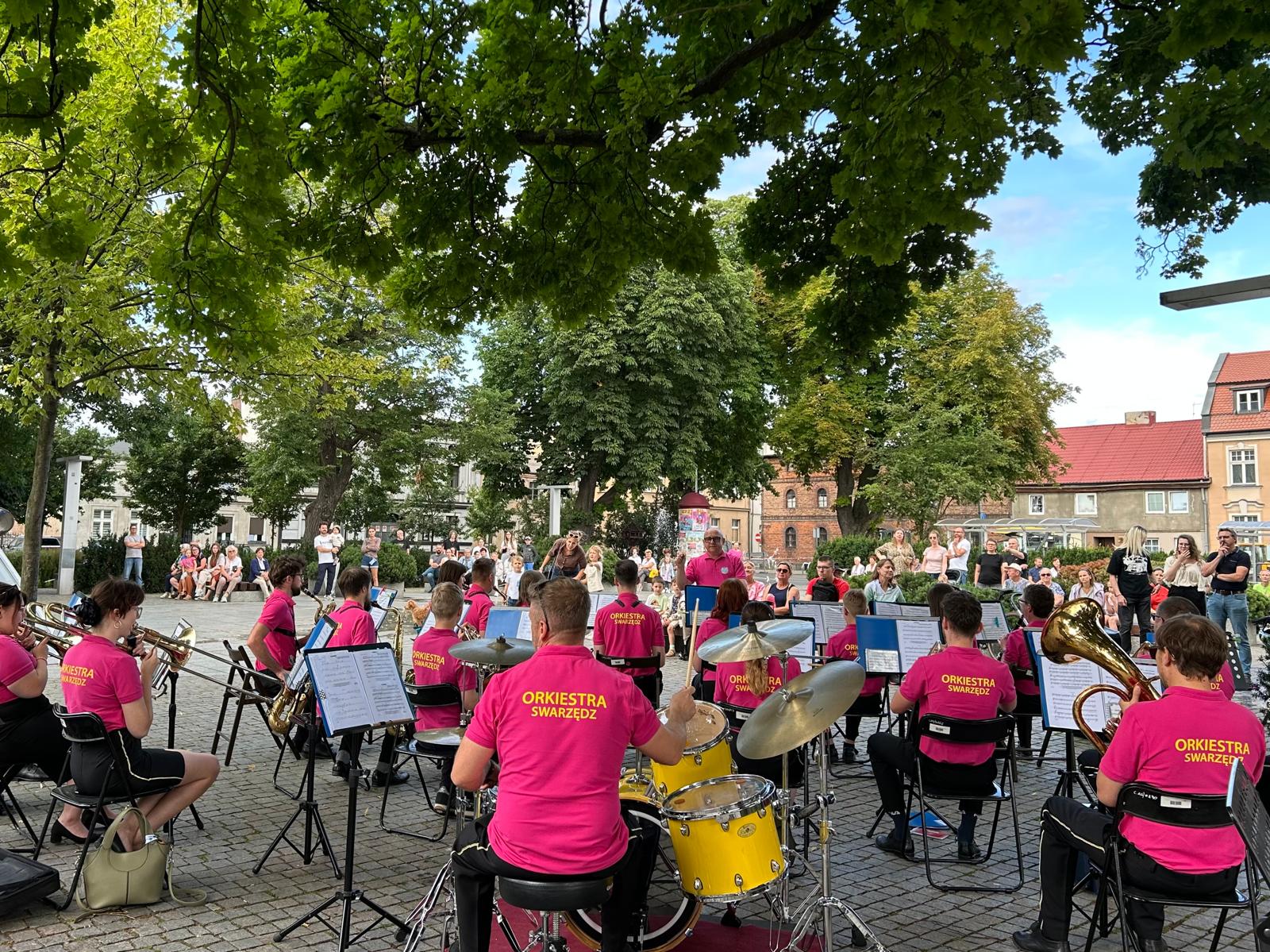Widok Placu Niezłomnych w Swarzędzu od strony orkiestry dętej wykonującej utwór. Orkiestranci siedzą na krzesłach, przed nimi stoi i dyryguje Łukasz Gowarzewski. W tle widoczna publiczność siedząca na ławkach i słuchająca muzyki, drzewa i kamienice.
