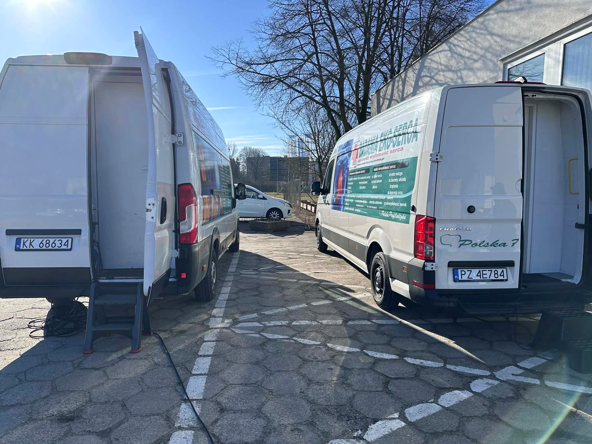 Zdjęcie przedstawia dwa samochody typu transporter służące do przeprowadzania badań serca i skóry stojące na parkingu przed Centrum Usług Społecznych.