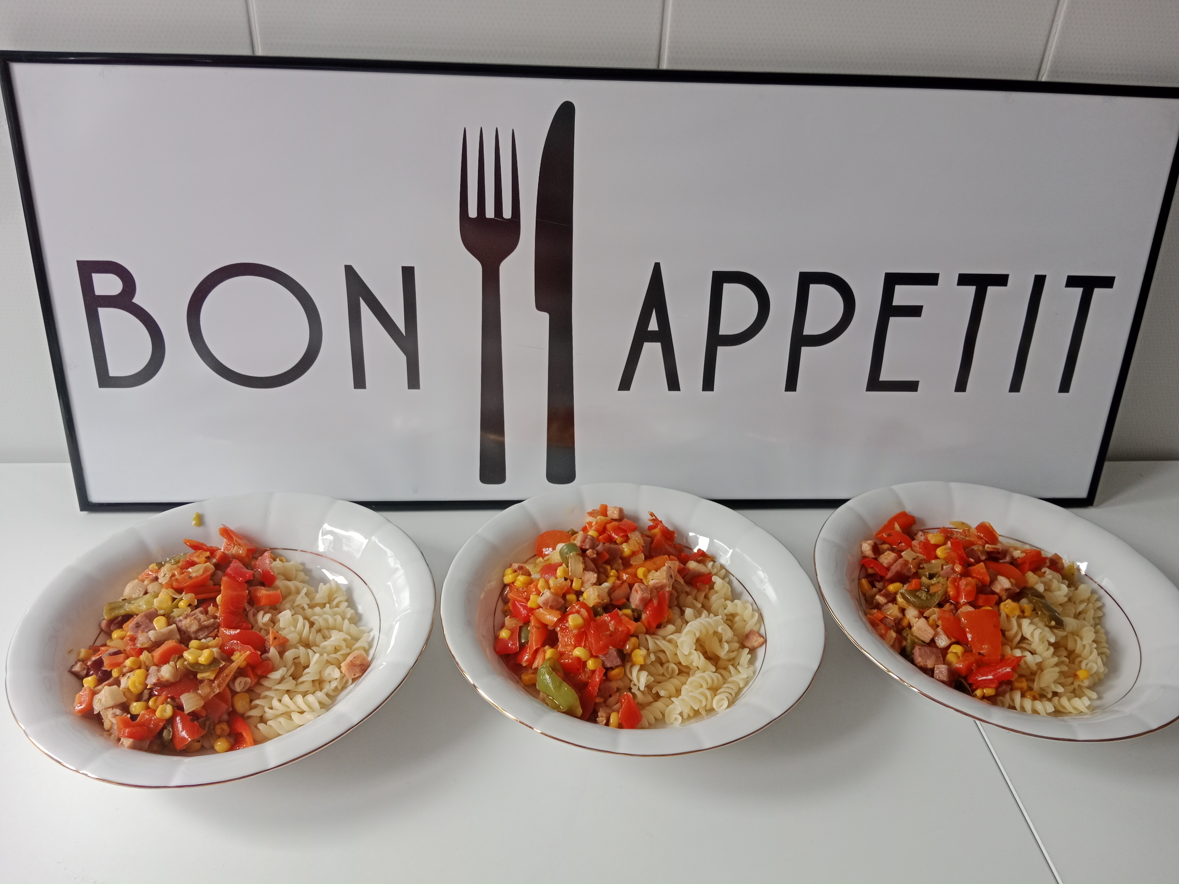 Na zdjęciu widać trzy białe głębokie talerze, w których jest makaron świderki, boczek i warzywa. Talerze stoją na tle tablicy z napisem Bon Apetit.