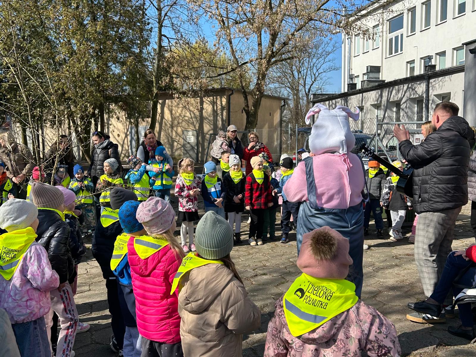 Ogród Społeczny. Grupa przedszkolaków z opiekunkami utworzyła krąg dokoła zająca maskotki i gitarzysty.