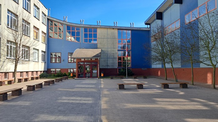 Czytnik e-booków Kindle na tle w pastelowych kolorach. Na ekranie widnieje czarny napis: „Młodzieżowy klub książki – wypożyczalnia Kindli”. W lewym górnym rogu fioletowy, rysunkowy kwiatek, a po prawej stronie strzałka w tym samym kolorze.