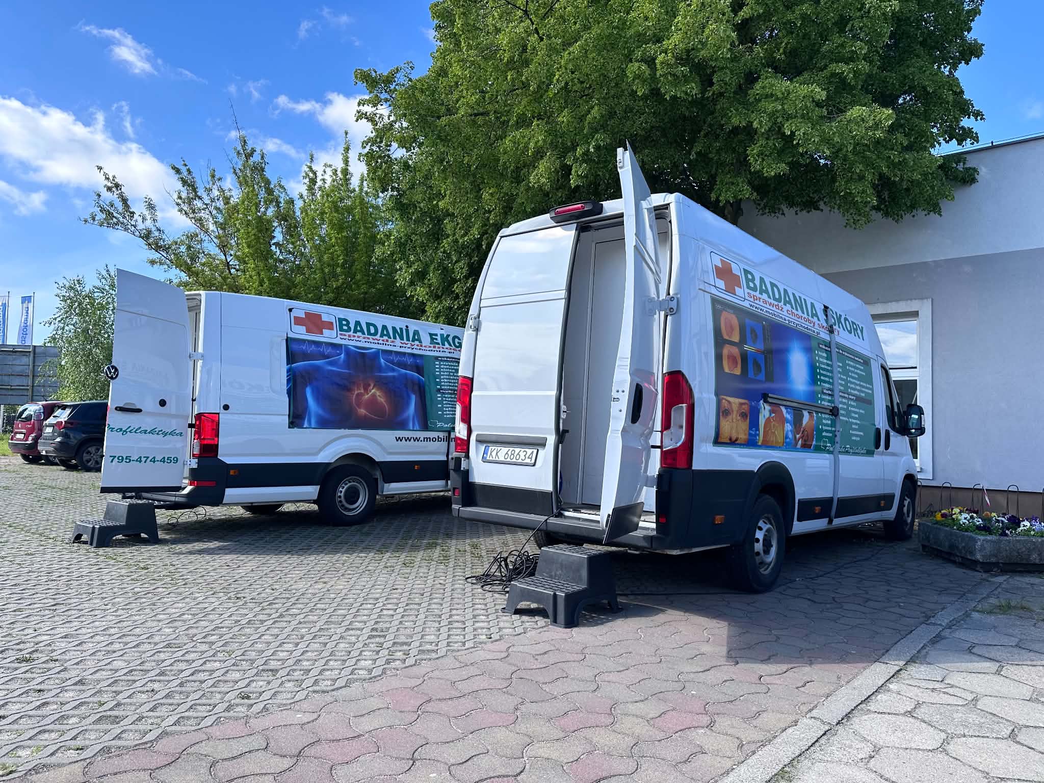 Na zdjęciu widać dwa duże busy diagnostyczne stojące na parkingu przy Centrum Usług Społecznych w Swarzędzu. W tle widać drzewa, błękit nieba i chmury.
