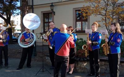 Zespół muzyczny w mundurach gra na instrumentach dętych na świeżym powietrzu, przed budynkiem w Swarzędzu. Na pierwszym planie widoczny jest dyrygent, a w tle baner z napisem 