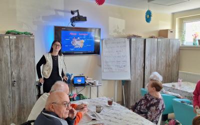 Zdjęcie przedstawia uśmiechniętą Żuliet stojącą frontem do seniorów siedzących przy stołach pokrytych ozdobną ceratą z motywami lawendy. Na ekranie widocznym za plecami wolontariuszki wyświetlony jest slajd poświęcony Alzacji i Lotaryngii.