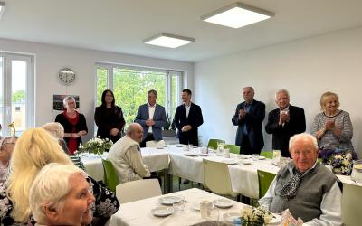Za uroczystymi stołami siedzą uśmiechnięci seniorzy i seniorki. W tle przy dwóch długich stołach stoją: dyrektor Centrum Usług Społecznych, pierwsza wicewojewoda, burmistrz i jego pierwszy zastępca, przewodnicząca rady miejskiej w Swarzędzu i radni.