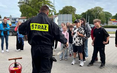 Zdjęcie przedstawia punkt startowy gry miejskiej pod tytułem Ratujemy bo umiemy. Na pierwszym planie widać strażaka tłumaczącego grupie uczniów konkurencję, do której za chwilę przystąpią. Obok strażaka stoi czerwona hydronetka wodna z żółtą etykietą.