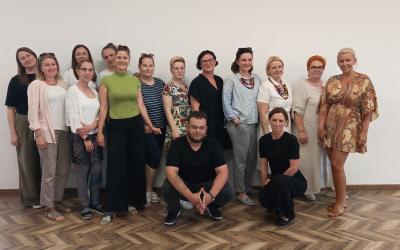 Zdjęcie wykonane w Centrum Międzypokoleniowym Pogodna. W sali na tle ściany stoi trzynaście przedstawicielek kół gospodyń wiejskich, kuca jeden mężczyzna również członek jednego z kół oraz organizatorka społeczności lokalnej.