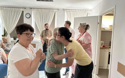Wnętrze Dziennego Domu Pomocy. Pożegnanie Żuljet Żama wolontariuszki Europejskiego Korpusu Solidarności. Wolontariuszka przytula się do seniorki składającej jej życzenia. Druga seniorka czeka w kolejce do Żuljet. W tle widać troje pracowników.