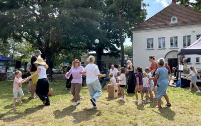 Zdjęcie wykonano na trawie przy budynku Ośrodka Kultury w Swarzędzu. Na pierwszym planie widoczne radośnie tańczące kobiety z dziećmi. W tle orkiestra flażoletowa, namioty z przekąskami i wysokie drzewa.