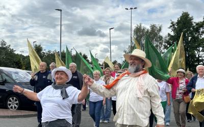 Senioriada. Korowód seniorów przechodzi przez parking przed pływalnią Wodny Raj. W pierwszej parze idzie radna Rady Miejskiej Seniorów i wodzirej.