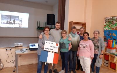 Wnętrze Centrum Aktywności Seniora. Wolontariusz europejski stoi w otoczeniu uśmiechniętych seniorek. Jedna z nich trzyma flagę Francji i transparent.