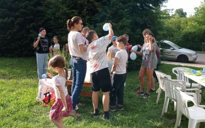Zdjęcie podczas zajęć kreatywnych na świeżym powietrzu. Pani instruktorka oraz grupka dzieci w wieku szkolnym z balonami w rękach.