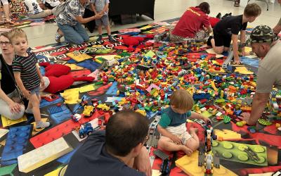 Piknik Trzech Pokoleń. Wnętrze namiotu przed sceną nad jeziorem. Na pierwszym planie widać rodziców z dziećmi bawiących się klockami lego, w tle widać kolejne stanowisko z klockami lego i sztalugi do malowania intuicyjnego.