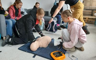 Gra miejska pod tytułem Ratujemy, bo umiemy. Na podłodze klęczą dwie dziewczynki. Jedna z nich układa fantoma. Druga trzyma w rękach elektrody AED.