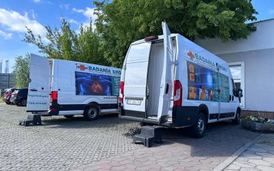 Na zdjęciu widać dwa duże busy diagnostyczne stojące na parkingu przy Centrum Usług Społecznych w Swarzędzu. W tle widać drzewa, błękit nieba i chmury.