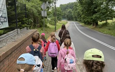 Widok zza uczestników półkolonii na szosę biegnącą przez łąki i las. Grupa prowadzona przez wychowawczynię idzie po chodniku wzdłuż szosy.