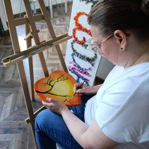Zdjęcie przedstawia uczestniczkę zajęć malującą pędzelkiem detale na płótnie w formie serca. Obrazek przedstawia czarne gałęzie na tle słońca.
