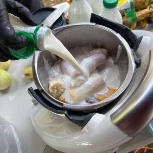 Zdjęcie przedstawia zbliżenie na garnek, w którym znajdują się banany, mango i inne owoce i do którego jest wlewany kefir.