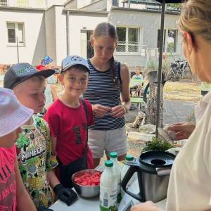 Zdjęcie przedstawia prowadzącą warsztaty kulinarne tłumaczącą jakąś kwestię dwóm dziewczynkom i dwóm chłopcom. W tle widać budynek Centrum Usług Społecznych 