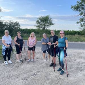 Grupa sześciu kobiet z kijkami podczas treningu nordic walking na terenie wiejskim. Uśmiechaja się do zdjęcia.