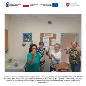 Zajęcia ruchowe w świetlicy wiejskiej w Paczkowie. Cztery uśmiechnięte seniorki na stojąco wykonują ćwiczenie ruchowe dłoni i ramion.