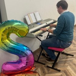 Chłopak gra na keyboardzie, siedzi na małym stołku z czerwonym obiciem. Po lewej stronie stoi nadmuchany balon w kształcie cyfry 2.