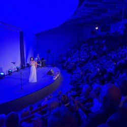 Swarzędzka Sala Koncertowa oświetlona niebieskim światłem. Na scenie występuje solistka Marzena Kurpisz i akompaniator Krzysztof Gruszka.