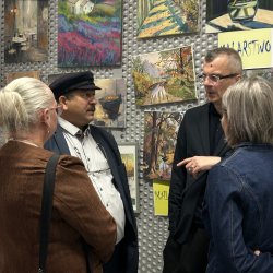 Rozmowa w kuluarach. Dwie kobiety i dwóch mężczyzn rozmawia ze sobą na tle prac plastycznych seniorów. Jednym z rozmówców jest Rom w kaszkiecie na głowie.