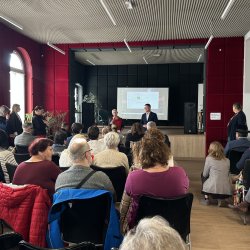 Widok ogólny na publiczność, słuchającą dyrektor Centrum Usług Społecznych. W tle scena i ekran z wyświetlonym slajdem.