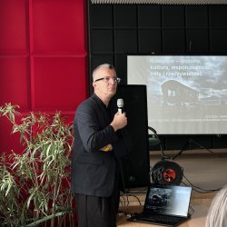 Jeden z prelegentów stoi przed sceną zwrócony w stronę publiczności, w ręce trzyma mikrofon. W tle widać ekran z wyświetlonym slajdem.