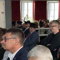 Widok uczestników konferencji siedzących na widowni w sali z lustrami w Centrum Aktywności Seniora. Wśród nich widać Roma w kapeluszu i kaszkiecie.