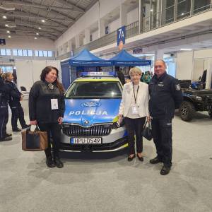 Wnętrze hali targowej. Jedna z laureatek w towarzystwie koordynatorki z Centrum Aktywności Seniora i policjanta stoi przy radiowozie.