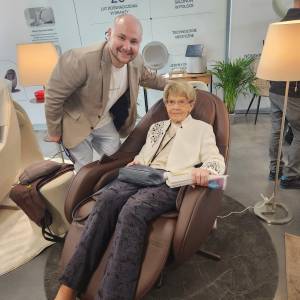 Wnętrze hali. Laureatka konkursu viva seniorzy siedzi na brązowym, regulowanym fotelu, który ma uniesiony podnóżek. Obok stoi uśmiechnięty instruktor.