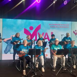 Zespół składający się z dwóch seniorów i ośmiu seniorek wykonuje na scenie utwór grając na ukulele. W tle widać kolorowe tło z napisem viva seniorzy.