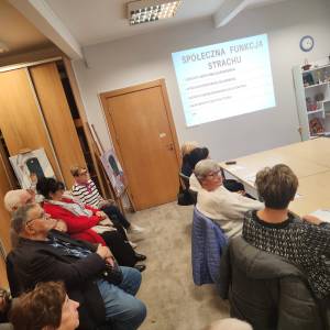 Zdjęcie przedstawia wnętrze sali w Centrum Aktywności Seniora, uczestników spotkania i ścianę z wyświetlonym slajdem.