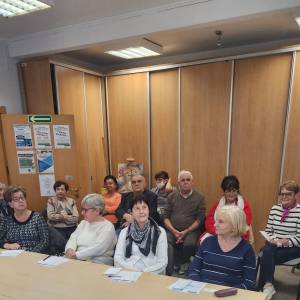 Zdjęcie przedstawia dwa rzędy seniorów siedzących na krzesłach i słuchających doktora Suworkina. Tło stanowi szafa przesuwana i otwarte drzwi do sali.  