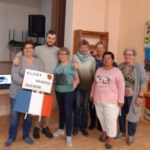 Wnętrze Centrum Aktywności Seniora. Wolontariusz z Francji Roma stoi w otoczeniu sześciu uśmiechniętych seniorek. Jedna z nich trzyma flagę Francji.