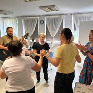 Zdjęcie przedstawia wnętrze Dziennego Domu Pomocy. Pracownik gra na gitarze, seniorki w kółeczku tańczą z Żuljet, wolontariuszką z Francji.