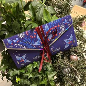 Prezent w postaci pięknej, świątecznej koperty z czerwoną kokardą na tle zielonych liści bluszczu i gałęzi świerku przystrojonych bombkami.