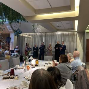 Wigilia w Eurohotelu. Widok na seniorów siedzących przy stole i klaszczących. W tle widać dyrekcję Centrum, księdza i władze miasta.